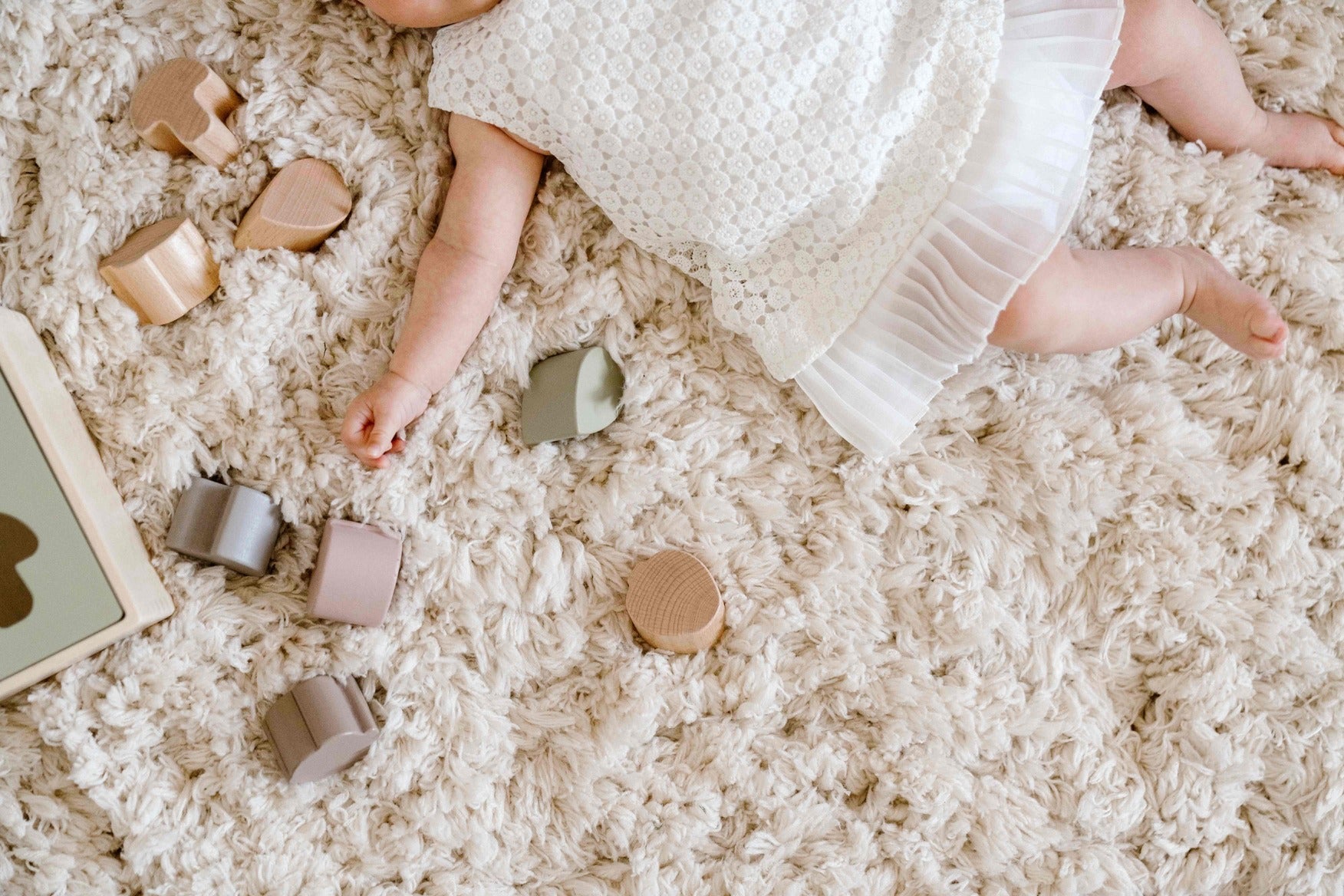 Baby liggend op een zacht, hoogpolig crèmekleurig vloerkleed, omringd door houten blokjes en een vormensorteerdoos in een rustige, minimalistische sfeer