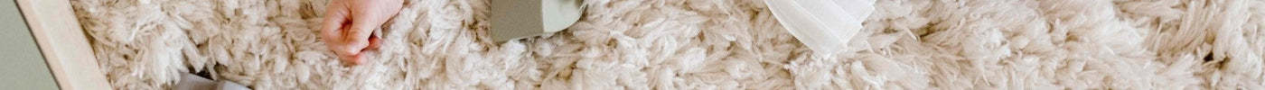 Baby liggend op een zacht, hoogpolig crèmekleurig vloerkleed, omringd door houten blokjes en een vormensorteerdoos in een rustige, minimalistische sfeer